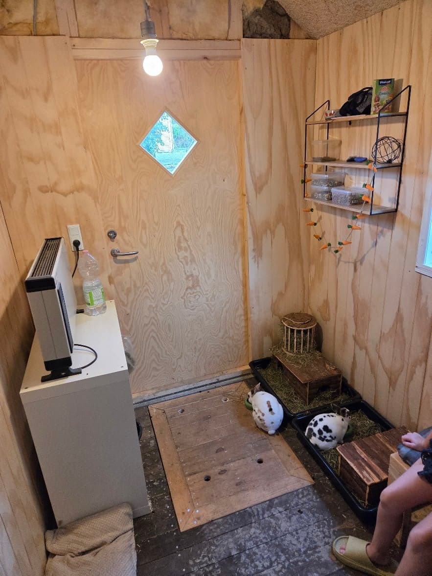 Kaninhuset indefra med døren, en hylde med foder, en varmeovn og kaninerne i deres bakker